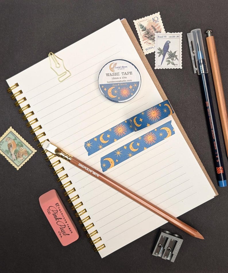 May include: A stationery flat lay featuring a spiral-bound notebook, washi tape, pencils, stamps, and an eraser. The washi tape has a blue background with a sun, moon, and star pattern. The notebook is open to a lined page.