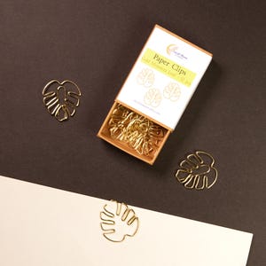 May include: Gold-colored Monstera leaf paper clips scattered on a brown and white surface. A box of paper clips is open, revealing more of the same design. The box reads "Paper Clips Gold Monstera Leaf - 12 pcs."