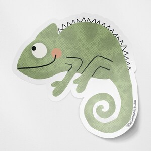 A photo of a die-cut glossy vinyl sticker featuring a cute hand-drawn green chameleon smiling.