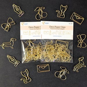 May include: Two clear plastic packages of gold-toned metal paper clips labeled Deco Paper Clips 12 pcs. The clips are shaped like envelopes, fountain pen nibs, dachshunds, cats, cherries, and bows, with loose clips scattered around the packages.