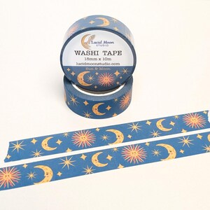 May include: Two rolls of washi tape with a celestial design. The tape is navy blue with a pattern of yellow moons, suns, and stars. The label reads "Washi Tape" and "Sun & Moon". A strip of the tape is unrolled.