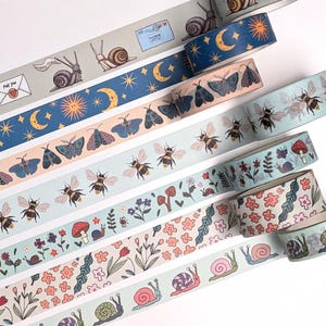 A collection of decorative washi tape rolls featuring various nature-inspired designs. The tapes showcase illustrations of snails, bees, butterflies, flowers, and celestial elements like moons and stars. The color palette includes soft blues, greens, and pinks.