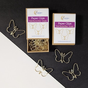 May include: Two rectangular kraft paper boxes labeled Paper Clips Gold Butterflies - 12 pcs. with the Lucid Moon Studio logo. One box is open, revealing gold butterfly-shaped wire paper clips inside. Four additional gold butterfly paper clips lie outside.