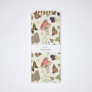 May include: A spiral-bound to-do notebook with a whimsical pattern of butterflies, mushrooms, snails, and berries. The notebook features the text "To-Do Notebook" on a white band. The design is printed on cream-colored paper.