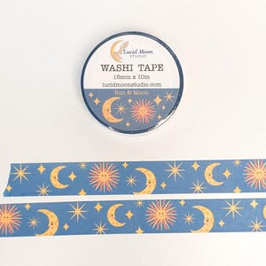 May include: A roll of washi tape with a celestial design featuring moons, suns, and stars on a dark blue background. The tape is 15mm x 10m. The text on the roll reads "Washi Tape" and "Sun & Moon".