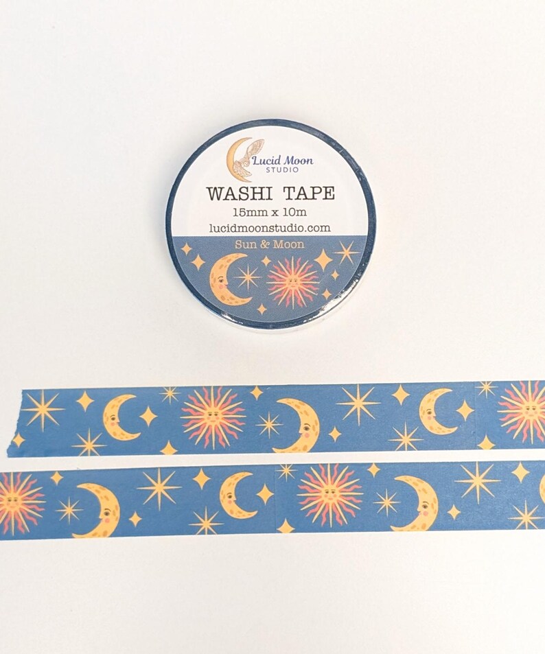 May include: A roll of washi tape with a celestial design featuring moons, suns, and stars on a dark blue background. The tape is 15mm x 10m. The text on the roll reads "Washi Tape" and "Sun & Moon".
