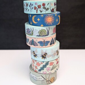 A stack of decorative washi tape rolls with various nature-themed designs. The rolls feature illustrations of flowers, mushrooms, bees, snails, moths, and celestial bodies in shades of blue, pink, and green. The bottom roll features snails holding a banner with the text "Special Delivery".