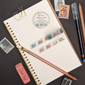A stationery flat lay featuring a spiral-bound notebook, washi tape with snail illustrations, stamps, pens, a pencil, eraser, pencil sharpener, and a gold snail paper clip. The washi tape is 15mm x 10m.