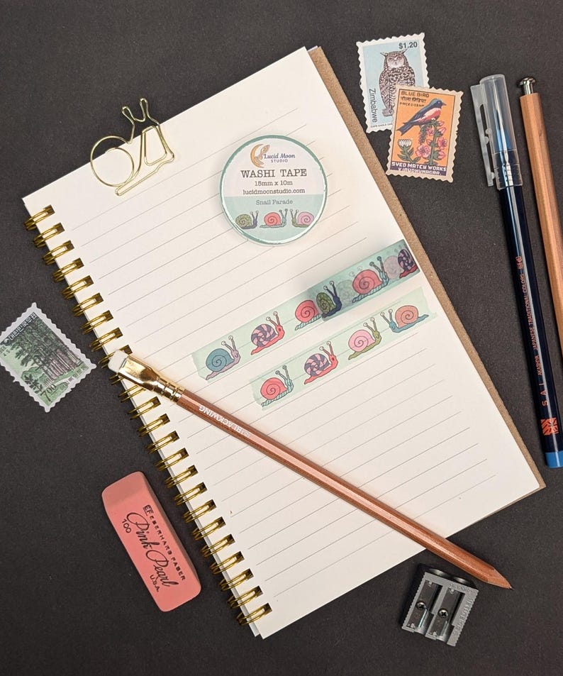A stationery flat lay featuring a spiral-bound notebook, washi tape with snail illustrations, stamps, pens, a pencil, eraser, pencil sharpener, and a gold snail paper clip. The washi tape is 15mm x 10m.