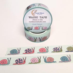 Two rolls of washi tape with a hand-drawn snail pattern. The tape is light aqua with colorful snails. The text on the tape reads "Washi Tape", "15mm x 10m", and "Snail Parade". The Lucid Moon Studio logo is also visible.