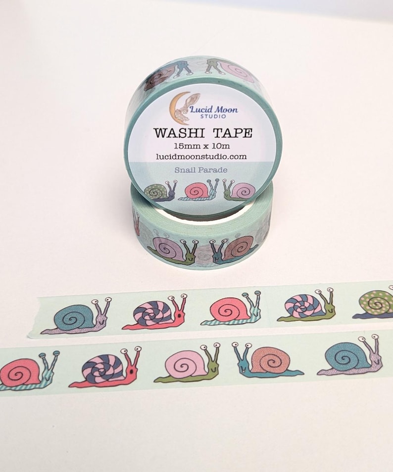 Two rolls of washi tape with a hand-drawn snail pattern. The tape is light aqua with colorful snails. The text on the tape reads "Washi Tape", "15mm x 10m", and "Snail Parade". The Lucid Moon Studio logo is also visible.