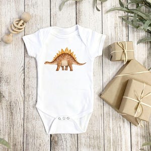 White baby bodysuit mockup with a brown watercolor stegosaurus dinosaur print, shown on rustic wood with wrapped gifts and greenery for kids apparel design.