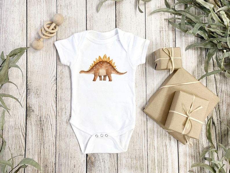 White baby bodysuit mockup with a brown watercolor stegosaurus dinosaur print, shown on rustic wood with wrapped gifts and greenery for kids apparel design.