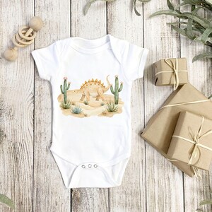 May include: White baby onesie with a watercolor illustration of a dinosaur in a desert landscape with cacti.