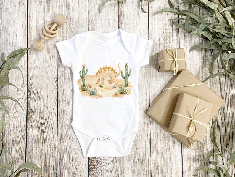 May include: White baby onesie with a watercolor illustration of a dinosaur in a desert landscape with cacti.