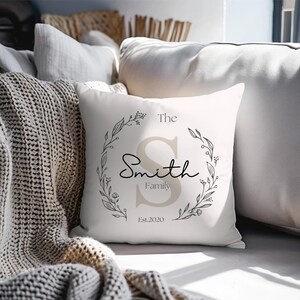 Puede incluir: Un cojín cuadrado blanco con un diseño de corona floral. El cojín presenta el texto "The Smith Family", una gran letra "S" y el año "Est. 2020". El diseño utiliza tonos grises y beige.