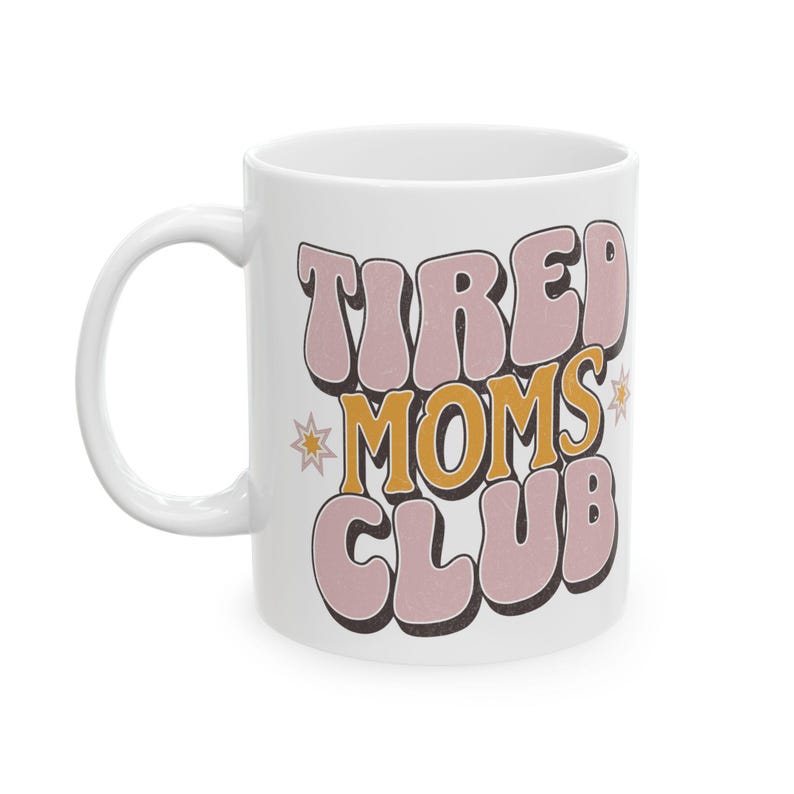 May include: White ceramic mug with the text "TIRED MOMS CLUB" in a retro font. The text is in pink and yellow with a black outline, and there are star accents. The mug has a curved handle.