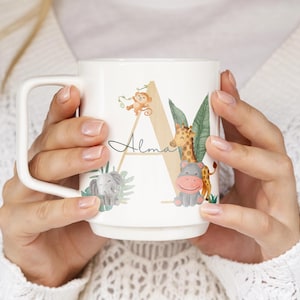 Puede incluir: Taza de cerámica blanca con un diseño de temática selvática. La taza muestra la letra "A" con una ilustración de jirafa, hipopótamo, mono y elefante. El nombre "Alma" está escrito en cursiva. La taza tiene un asa cuadrada.