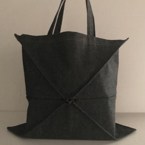 以下が含まれることがあります： デニムのような生地で作られた濃いグレーのトートバッグ。バッグは幾何学模様になるように折り畳まれています。バッグには2つのハンドルが付いています。