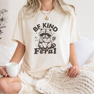 以下が含まれることがあります： 黒い文字で「BE KIND OF FERAL」と書かれた、花を持っているアライグマのグラフィックが特徴のクリーム色のTシャツ。アライグマは黒いマスクとふわふわの尻尾をしています。シャツは半袖でゆったりとしたフィット感です。