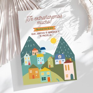 Peut inclure: Une carte de vœux avec le texte "¡Te extrañaremos mucho!" et "Numeros 6:24". La carte présente une illustration colorée de maisons nichées parmi des montagnes vertes sous un soleil jaune. La carte est sur une surface blanche.
