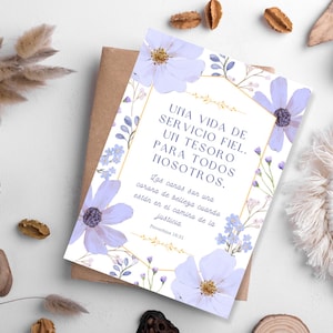 Può includere: Un biglietto di auguri con un design floreale con fiori lavanda e dettagli dorati. La carta ha testo in spagnolo. La carta è posizionata su una superficie bianca con elementi decorativi.