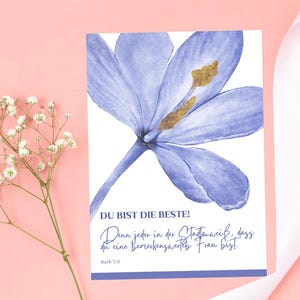 May include: A greeting card with a watercolor illustration of a blue flower with gold accents. The card has text in German that reads "DU BIST DIE BESTE!" and a quote. A sprig of white flowers and a white ribbon are on a pink background.