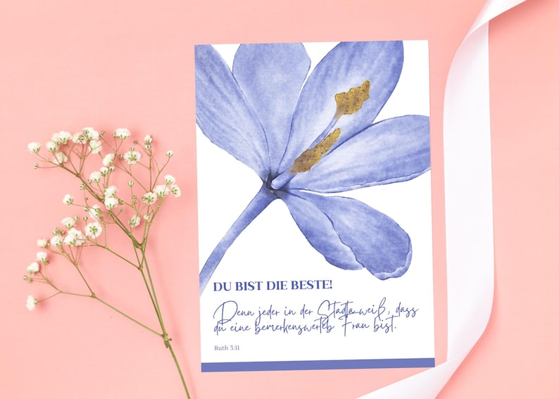 May include: A greeting card with a watercolor illustration of a blue flower with gold accents. The card has text in German that reads "DU BIST DIE BESTE!" and a quote. A sprig of white flowers and a white ribbon are on a pink background.