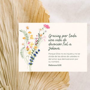 Può includere: Una carta color crema con una composizione floreale ad acquerello e testo in spagnolo. I fiori includono fiori rosa, gialli, blu e bianchi. Il testo recita: "Gracias por toda una vida de devoción fiel a Jehová."