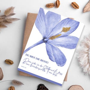 May include: A greeting card featuring a watercolor illustration of a large, light purple flower with yellow stamens. The card has text in German that reads "DU BIST DIE BESTE!" and is resting on a brown envelope.