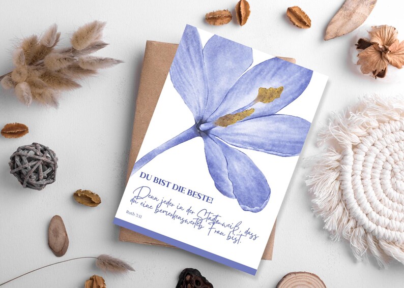 May include: A greeting card featuring a watercolor illustration of a large, light purple flower with yellow stamens. The card has text in German that reads "DU BIST DIE BESTE!" and is resting on a brown envelope.