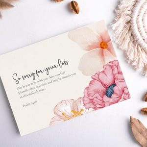 Puede incluir: Tarjeta de condolencia con ilustraciones florales en acuarela en tonos rosa y melocotón. La tarjeta presenta el texto "So sorry for your loss" y un mensaje reconfortante del Salmo 14:18. Elementos decorativos: posavasos de macramé y vainas secas.
