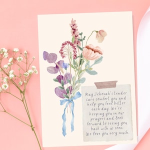 Op de afbeelding: Een wenskaart met een aquarel bloemstuk in roze, paarse en blauwe tinten, vastgebonden met een blauw lint. De kaart bevat een handgeschreven bericht op een kleine, met plakband bevestigde notitie. Een takje witte bloemen en een wit lint zijn ook zichtbaar.