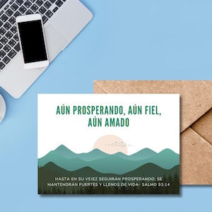 Può includere: Un biglietto di auguri con il testo "AÚN PROSPERANDO, AÚN FIEL, AÚN AMADO" su un'illustrazione di paesaggio di montagne e un sole al tramonto. Il biglietto è su una superficie blu con un laptop, un telefono, una tazza di caffè e una busta marrone.