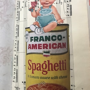 May include: A yellow can of Franco-American Spaghetti in tomato sauce with cheese. A baby wearing a blue and white outfit sits on top of the can, holding a pink toy.