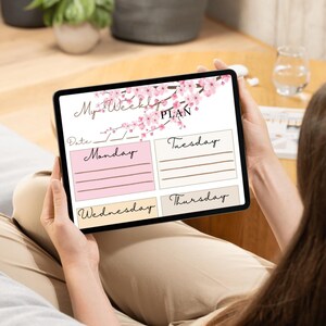 Peut inclure: Une tablette numérique affichant un planificateur hebdomadaire à l'écran. Le planificateur présente un motif floral rose et est intitulé "My Weekly PLAN". La tablette est tenue par une personne.