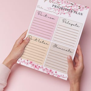 Op de afbeelding: Een notitieblok voor dagelijkse prioriteiten met een bloemmotief en vier secties: "Doe het nu", "Delegeer", "Doe het later" en "Elimineer". Het notitieblok wordt tegen een roze achtergrond gehouden.