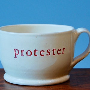 May include: White ceramic mug with a red inscription that reads "protester".