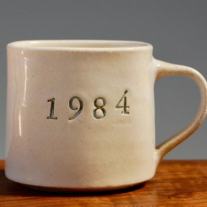 Könnte beinhalten: Eine cremefarbene Keramiktasse mit der Jahreszahl "1984" auf der Seite eingraviert. Die Tasse hat einen abgerundeten Henkel und eine leicht strukturierte Oberfläche. Die Tasse steht auf einer Holzoberfläche.