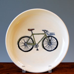 May include: A white ceramic plate with a green bicycle design. The bicycle has a brown seat and black wheels.