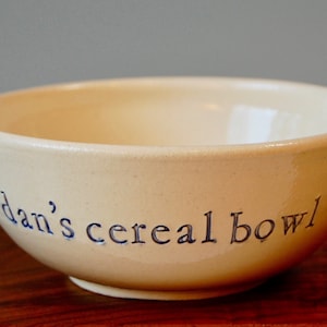 以下が含まれることがあります： クリーム色のセラミック製シリアルボウル。青い筆記体で「dan's cereal bowl」と書かれています。ボウルは丸みを帯びた形状で、滑らかで光沢のある仕上がりです。シンプルで日常的なキッチンアイテム。