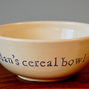 以下が含まれることがあります： クリーム色のセラミックボウルに、青い文字で「dan's cereal bowl」と書かれています。