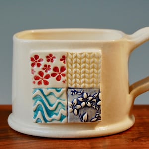 Pode incluir: Caneca de cerâmica branca com um design quadrado. A caneca tem quatro padrões diferentes: flores vermelhas e brancas, um padrão de malha amarelo, ondas azuis e verdes e flores azuis e brancas.