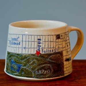Könnte beinhalten: Eine weiße Keramiktasse mit handgemaltem Design einer Stadtkarte. Die Karte zeigt einen roten Stern, der die Kreuzung der 8th Ave und der Fulton St. markiert. Die Tasse hat ein grünes und blaues Design um den Boden herum.