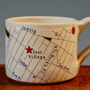 Può includere: Una tazza in ceramica bianca con una mappa del quartiere di East Village a New York City. La mappa include le strade "Bowery", "St. Mark's Pl.", "1st Ave.", "2nd Ave." e "Ave A". La tazza ha una stella rossa che segna la posizione di East Village.