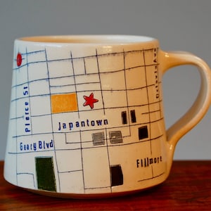 Könnte beinhalten: Weiße Keramiktasse mit einem Kartendesign mit den Worten "Japantown", "Geary Blvd", "Pierce St" und "Fillmore". Die Karte ist mit blauen und schwarzen Linien umrandet und enthält einen roten Stern und ein gelbes Rechteck.