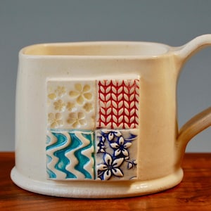 Pode incluir: Caneca de cerâmica branca com um design quadrado apresentando quatro padrões diferentes em azul, vermelho, branco e amarelo.