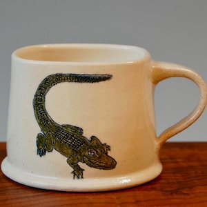 May include: A ceramic mug with a beige glaze and a green and black alligator illustration. The mug has a rounded body and a handle.