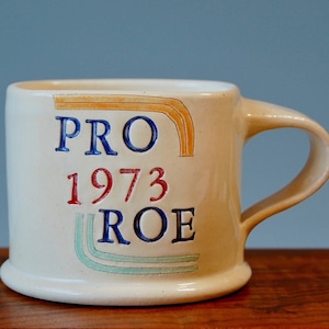 Puede incluir: Una taza de cerámica blanca con un diseño azul, rojo y naranja que dice "PRO 1973 ROE".