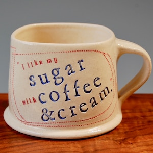 May include: A white ceramic coffee mug with a red and blue inscription that reads "I like my sugar with coffee & cream."
